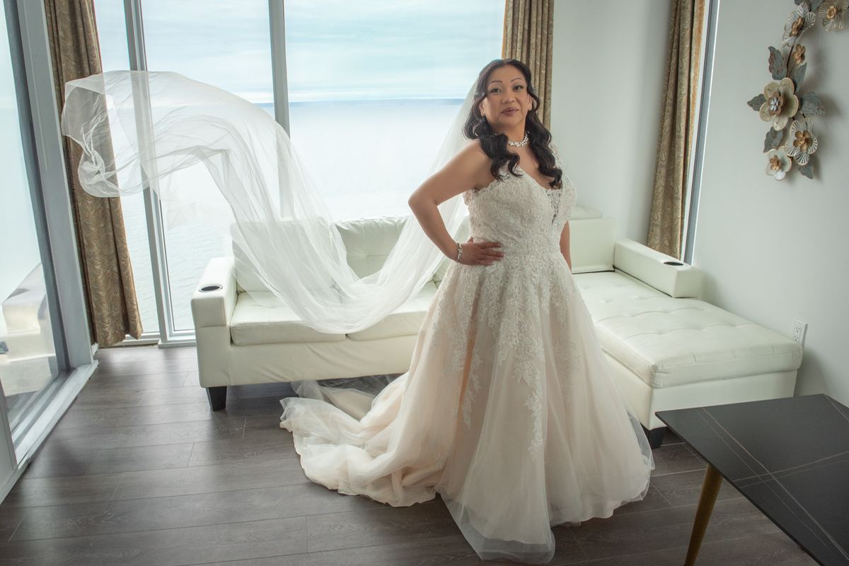 A bride in a lace wedding gown stands by windows overlooking the ocean, her veil fluttering in the breeze.