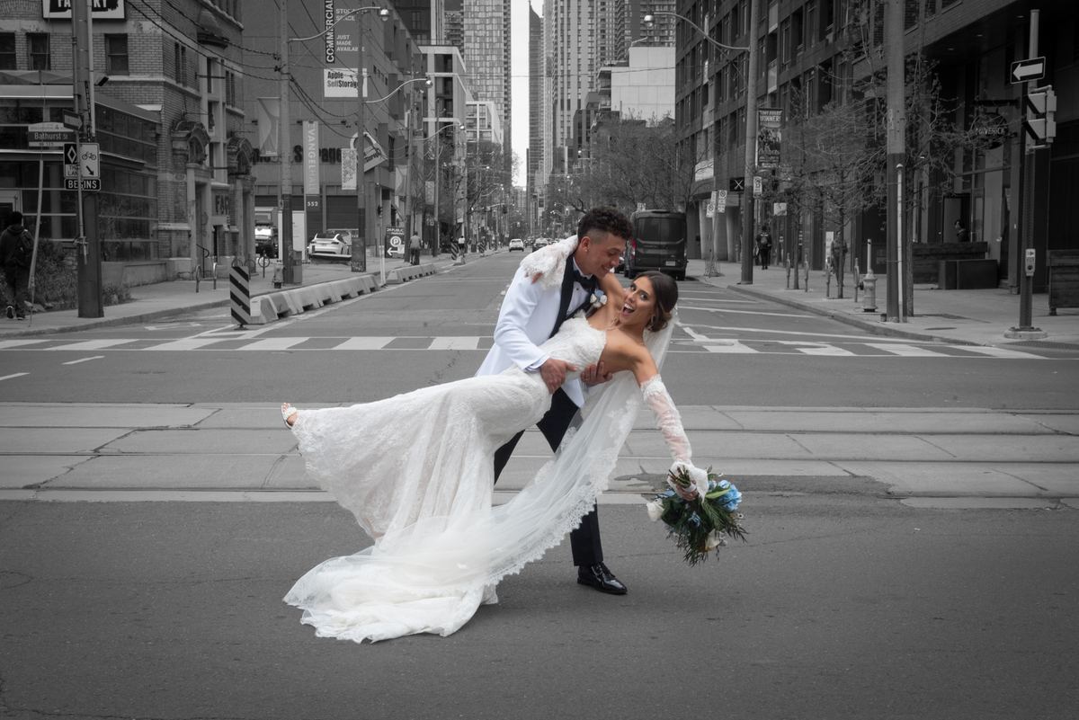 Bride laughing as groom lifts her on city street, dress billowing, bouquet colorful against black-and-white backdrop.