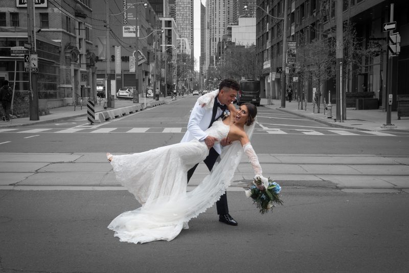 Bride laughing as groom lifts her on city street, dress billowing, bouquet colorful against black-and-white backdrop.