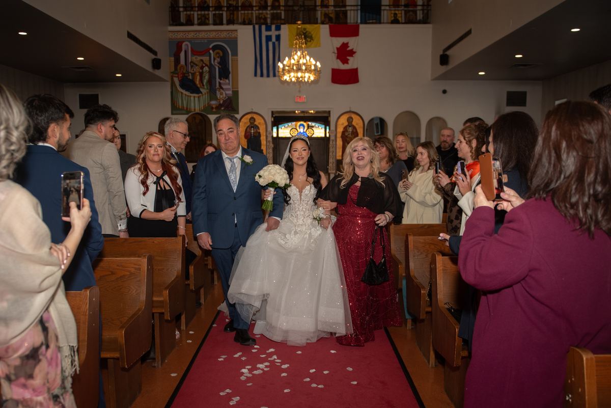 A bride in white walks down the aisle with her father and a woman in red as guests use phones, flags above altar visible.