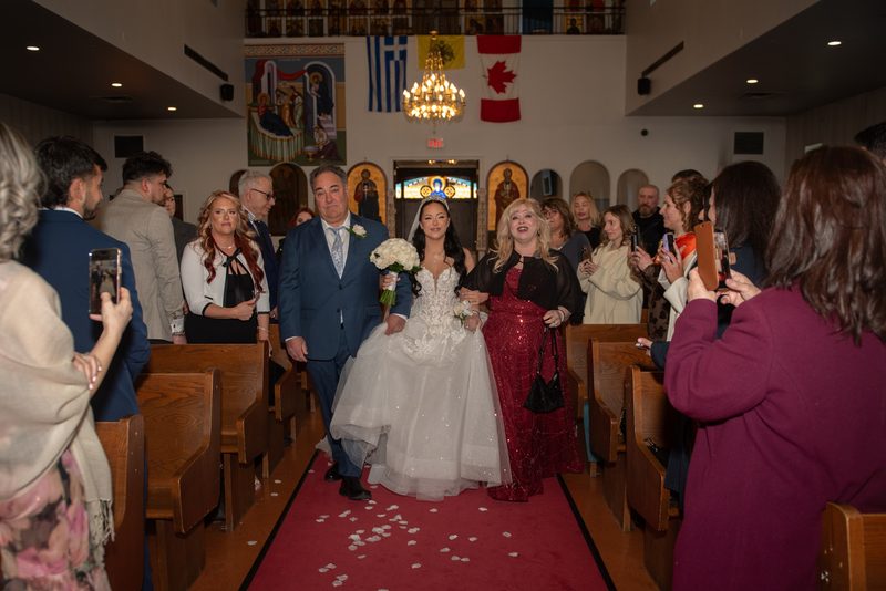 A bride in white walks down the aisle with her father and a woman in red as guests use phones, flags above altar visible.