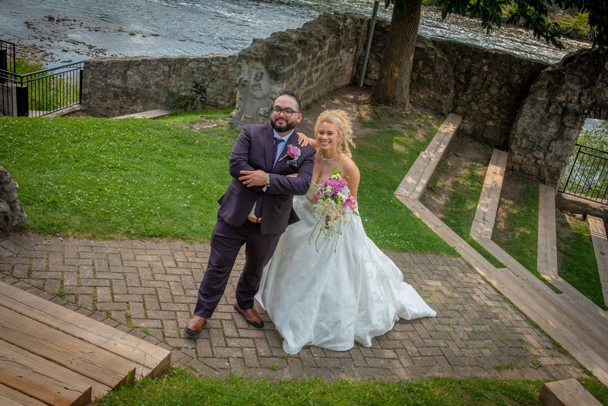 A smiling bride with a bouquet and her groom embrace on a stone walkway by water, surrounded by greenery.