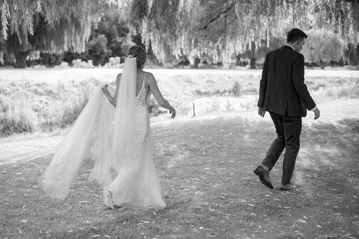 A bride and groom walk down a grassy path under a weeping willow, their figures silhouetted by dappled sunlight.