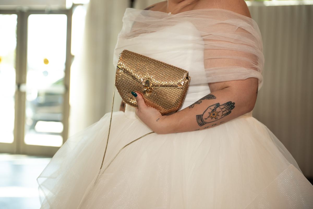 A bride in a voluminous white tulle gown holds a gold clutch, her arm featuring a tattoo of hands clasped around a star.