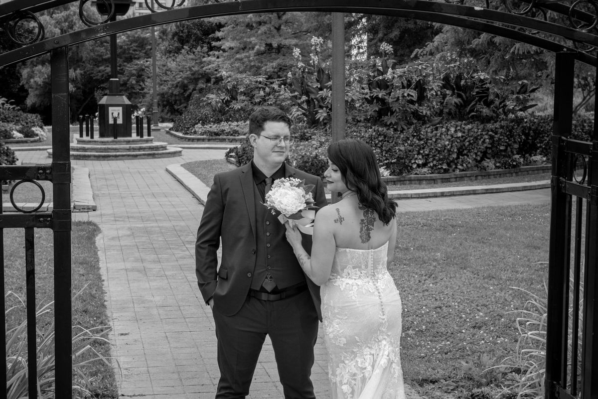 Bride in lace gown and groom in suit stand close by an ornate gate, gazing tenderly at each other in a garden.