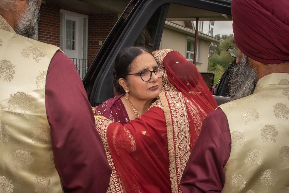 A bride in red and gold is helped into a car by two men in traditional Sikh attire, the scene filled with emotion.
