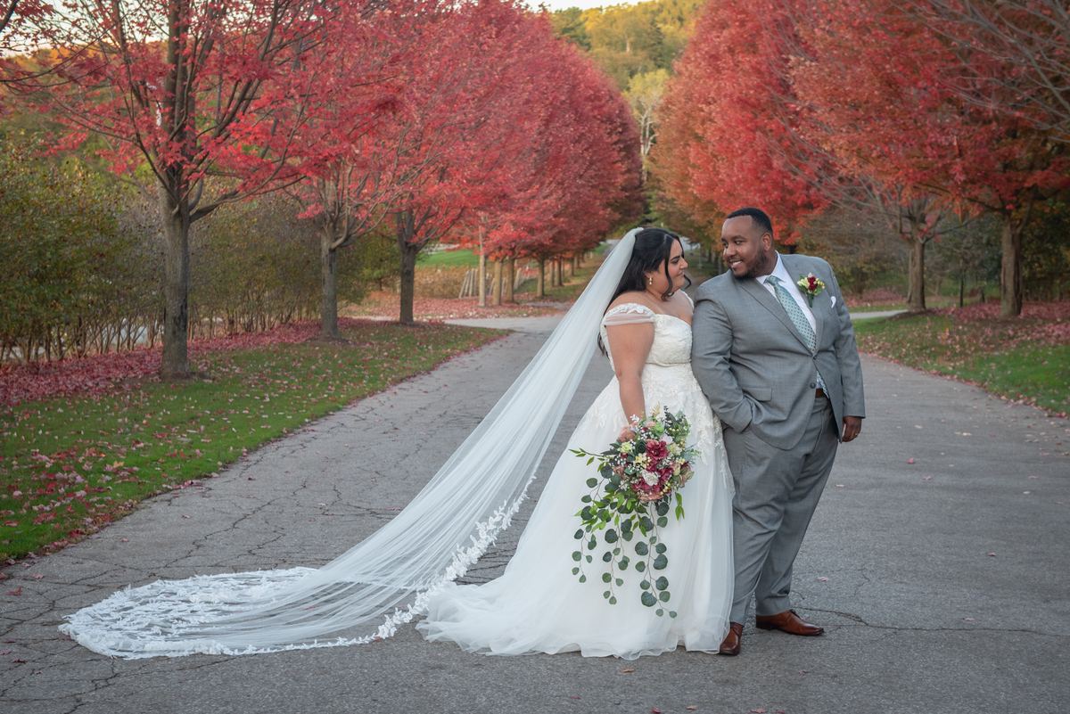 A newlywed couple stands closely on a path lined with crimson maple trees, bathed in golden sunlight.