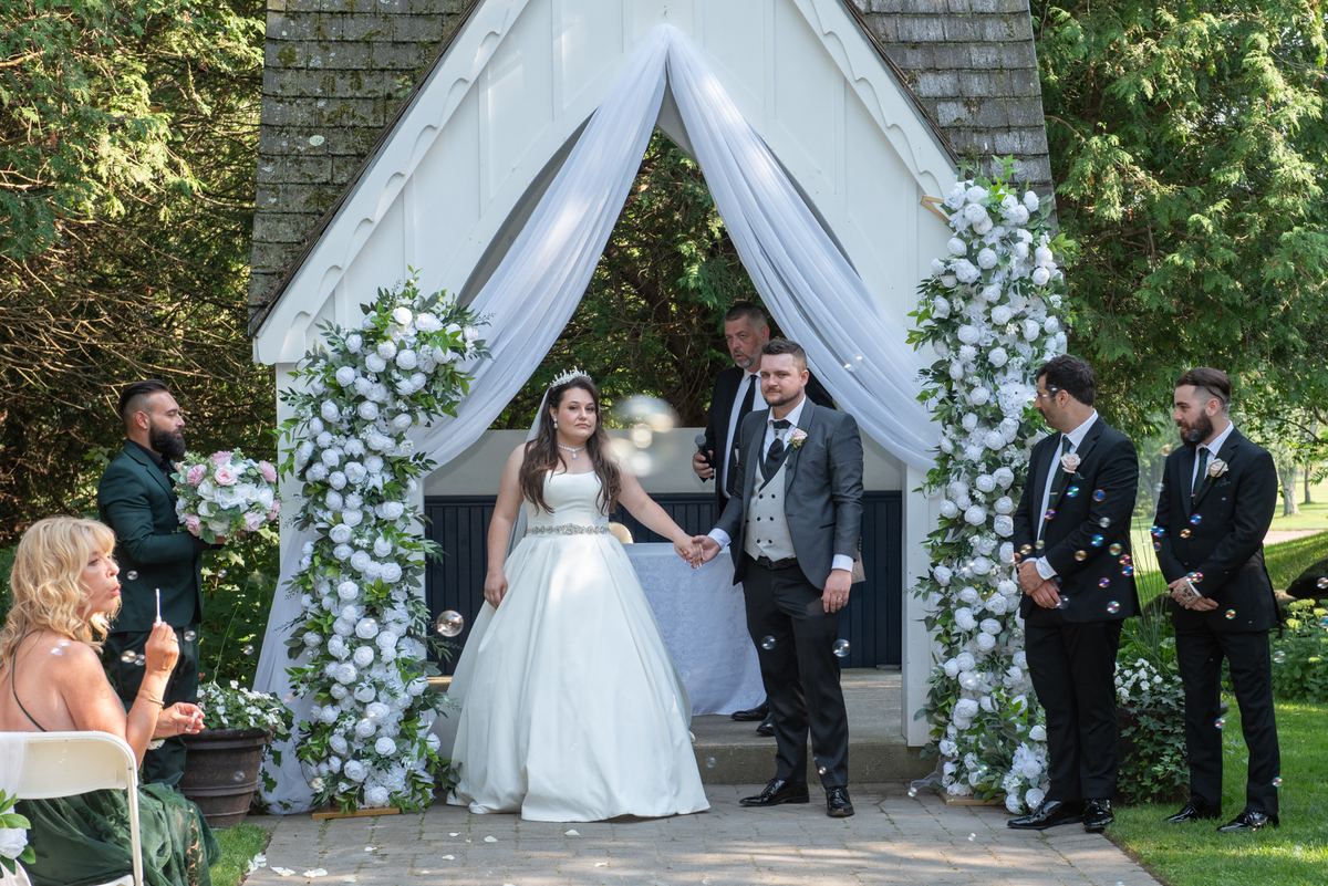 A bride and groom stand under a floral arch in a sunlit garden surrounded by guests and the bridal party.