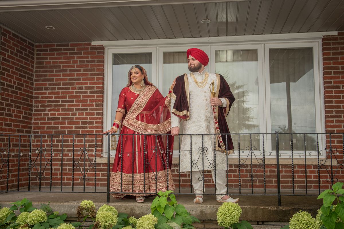 A South Asian bride and groom in traditional attire pose joyfully on a porch with a brick wall and wrought-iron railing.