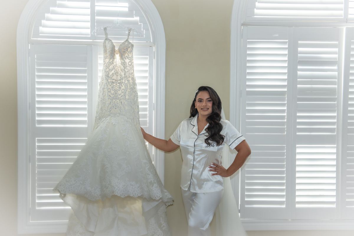 A bride in white satin pajamas with 'Bride' embroidered on the chest stands beside her wedding gown near an arched window.