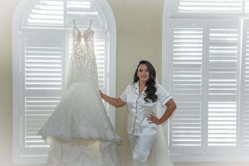 A bride in white satin pajamas with 'Bride' embroidered on the chest stands beside her wedding gown near an arched window.