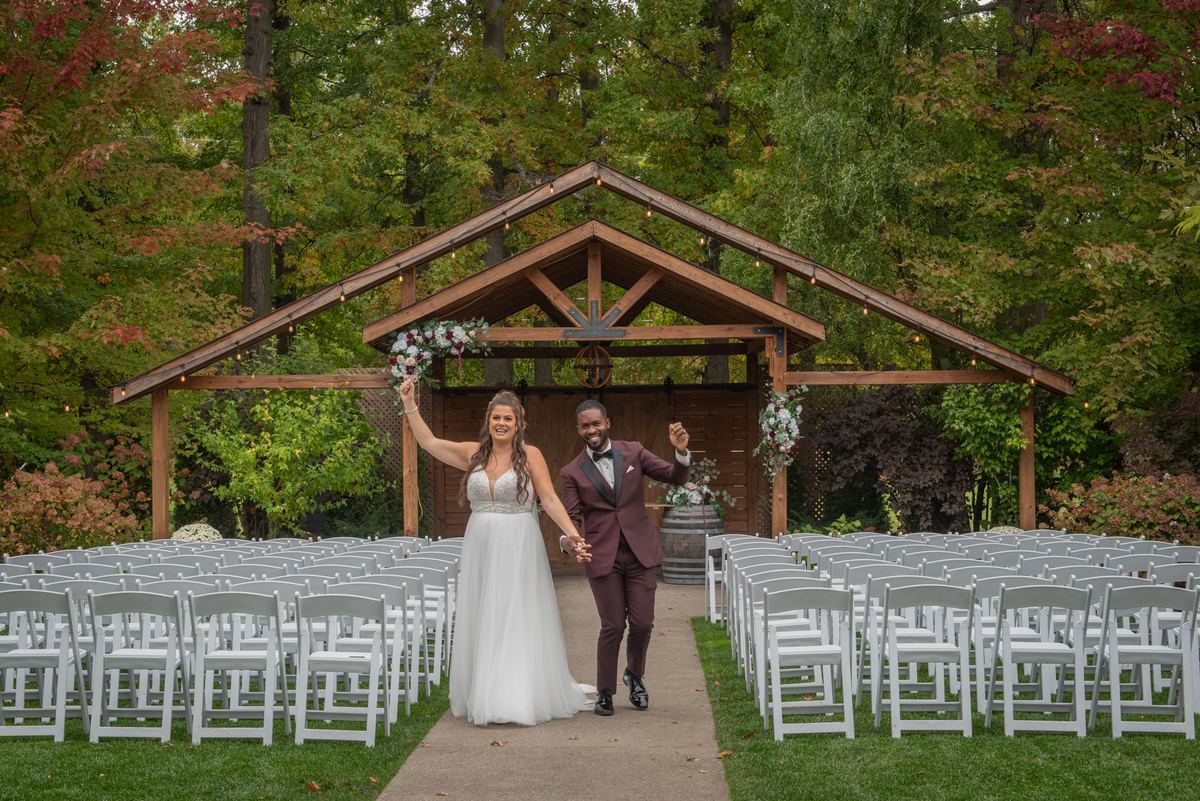 Bride and groom in formal attire celebrate their wedding outdoors under an arbor adorned with lights and flowers.