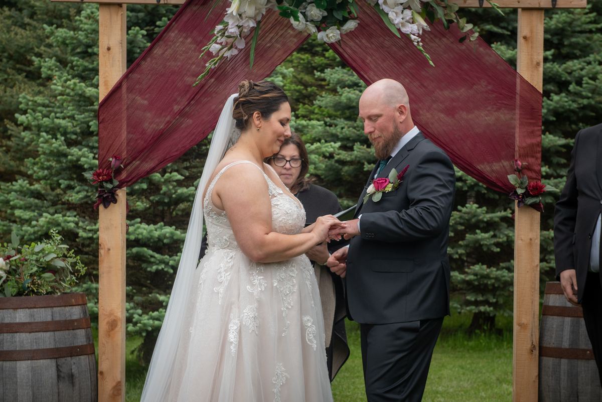 Bride and groom exchange rings under wooden arch adorned with burgundy fabric and flowers in an outdoor ceremony.