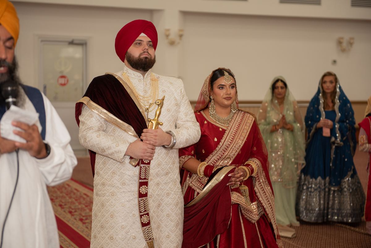 A Sikh groom in white and red stands beside his bride in a red and gold lehenga during a ceremony inside a gurdwara.