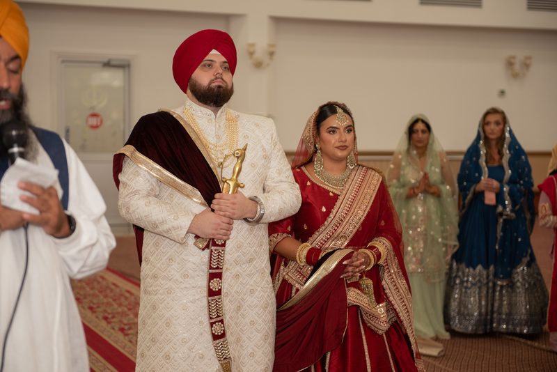 A Sikh groom in white and red stands beside his bride in a red and gold lehenga during a ceremony inside a gurdwara.
