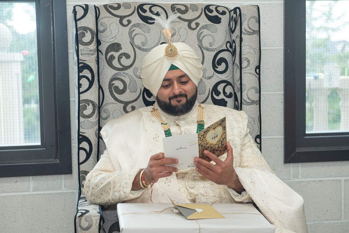 A groom in white ceremonial attire with a feathered turban reads a card while holding a decorated book.