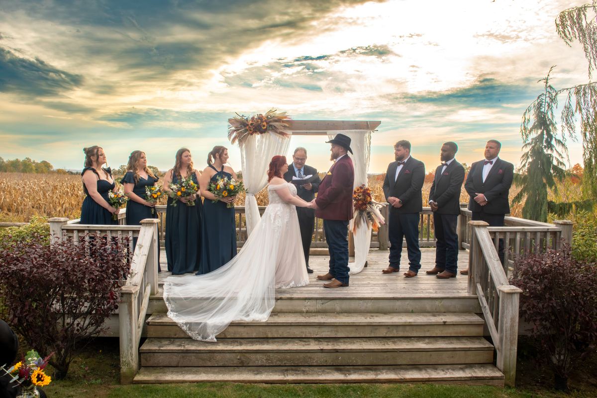 A bride and groom exchange vows under an autumnal floral arch on a wooden deck, surrounded by their wedding party.
