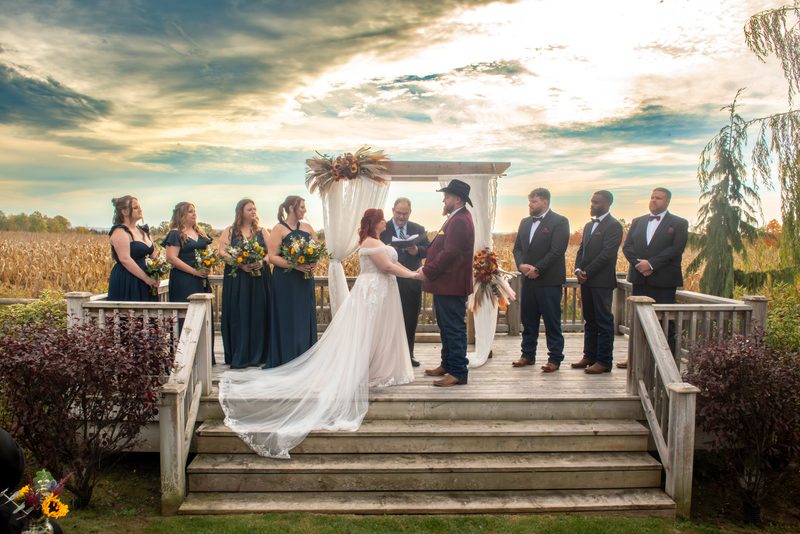 A bride and groom exchange vows under an autumnal floral arch on a wooden deck, surrounded by their wedding party.