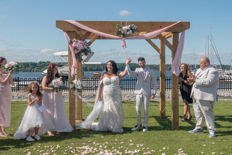 Bride and groom stand under wooden arch adorned with pink ribbons and flowers, surrounded by smiling guests.