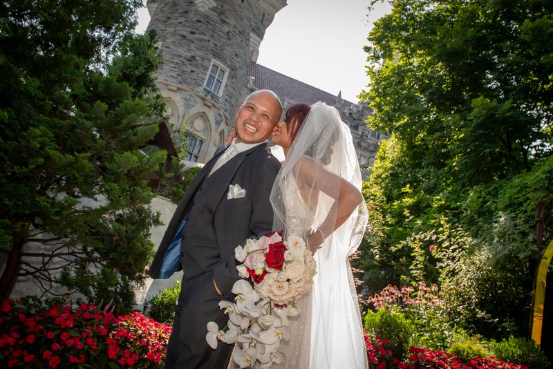A smiling groom and bride kiss, hand on shoulder, in front of a historic stone building under filtered sunlight.