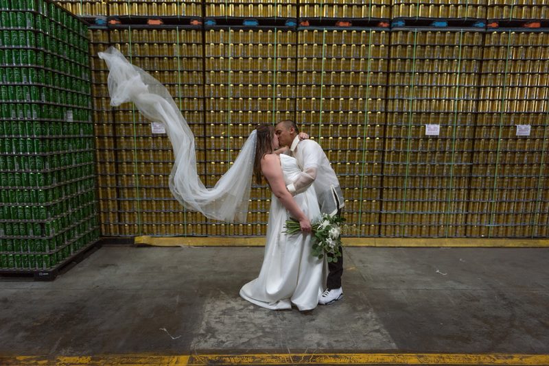 A bride and groom kiss in an industrial warehouse surrounded by stacks of green and gold beverage cans.
