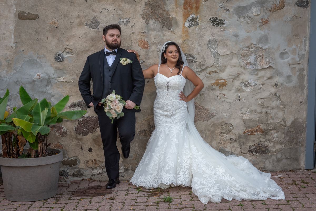 A newlywed couple in formal attire pose confidently against a weathered stone wall, illuminated by soft daylight.
