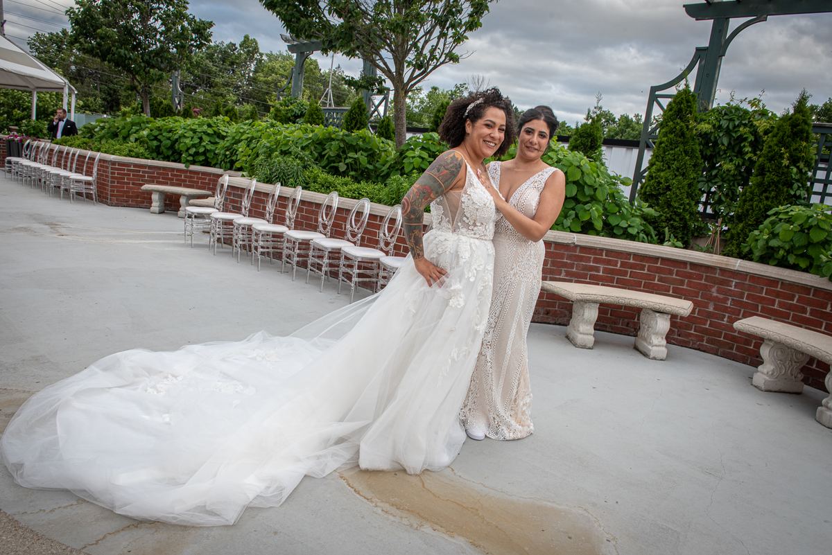 Two brides smile and embrace on a paved terrace beside rows of white chairs, framed by greenery and brick walls.