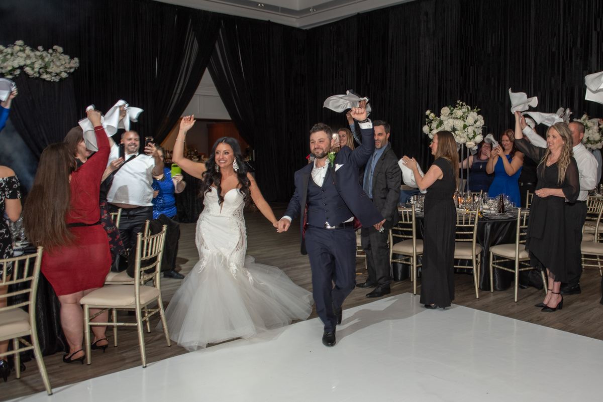 A bride and groom run down the aisle, guests waving white napkins in celebration at an elegant banquet hall.