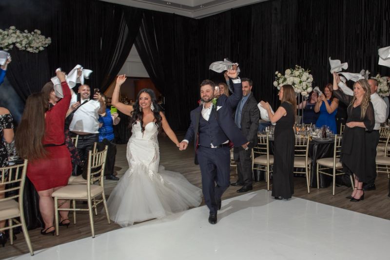 A bride and groom run down the aisle, guests waving white napkins in celebration at an elegant banquet hall.