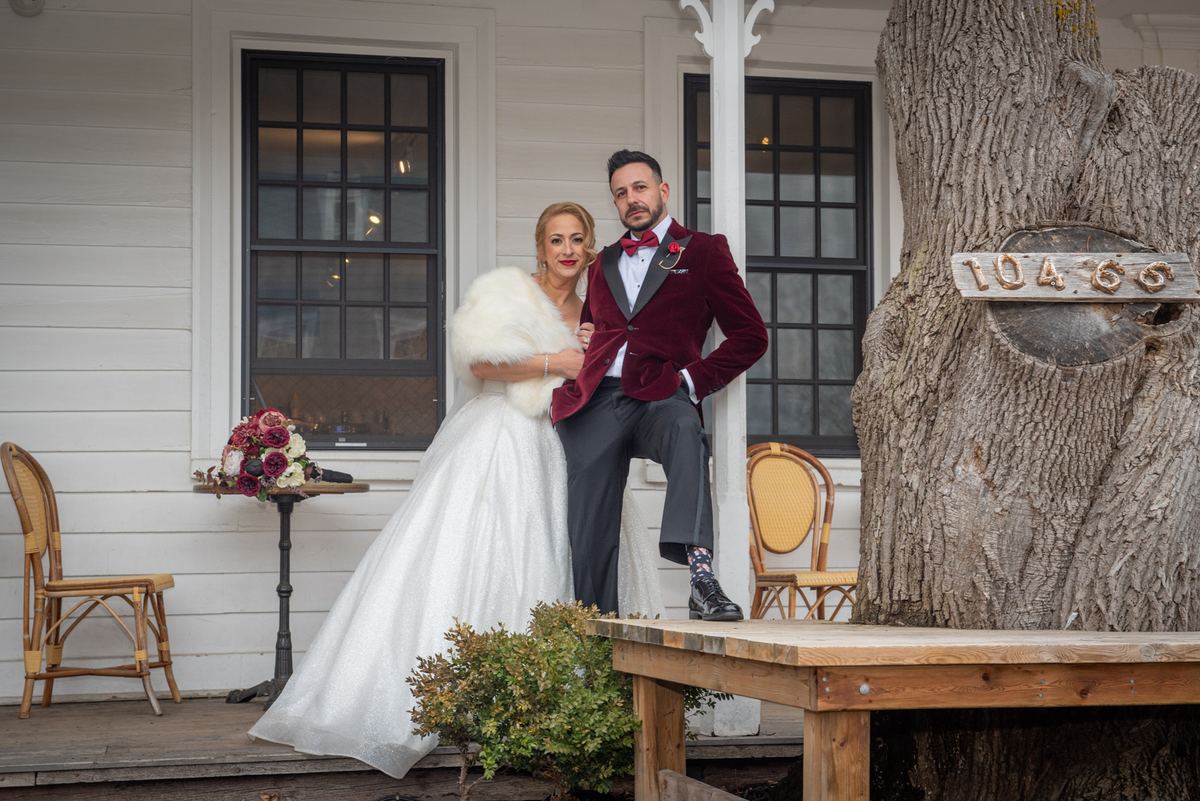 A bride in a white gown and fur stole stands with her groom in a burgundy tuxedo on a rustic porch beside a large tree.