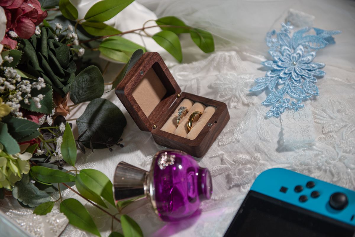 A wooden ring box open with two rings, beside flowers, lace appliqué, perfume bottle, and Nintendo Switch.