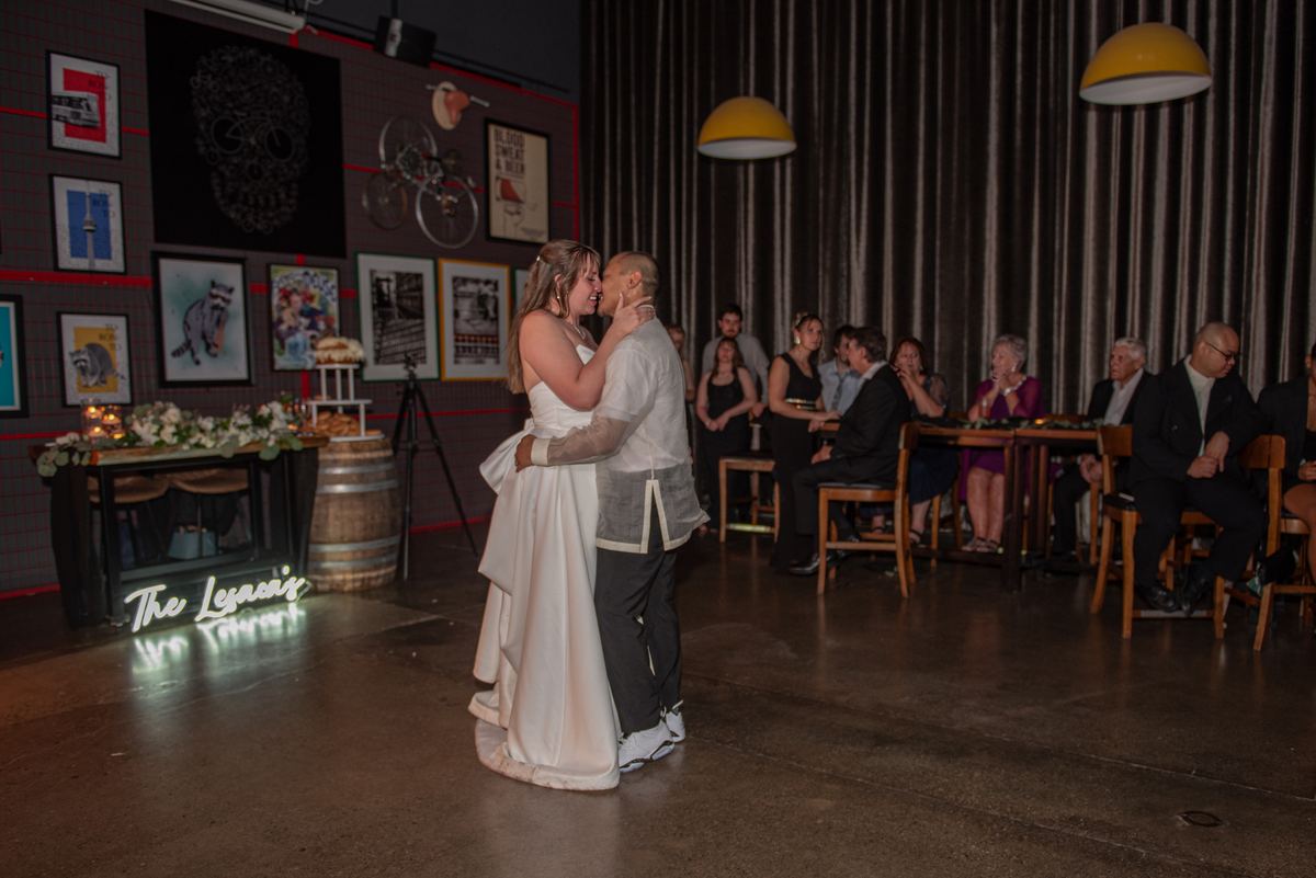 A bride and groom dance tenderly on a polished floor under hanging yellow lamps and a glowing neon sign.