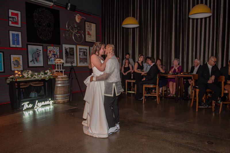 A bride and groom dance tenderly on a polished floor under hanging yellow lamps and a glowing neon sign.
