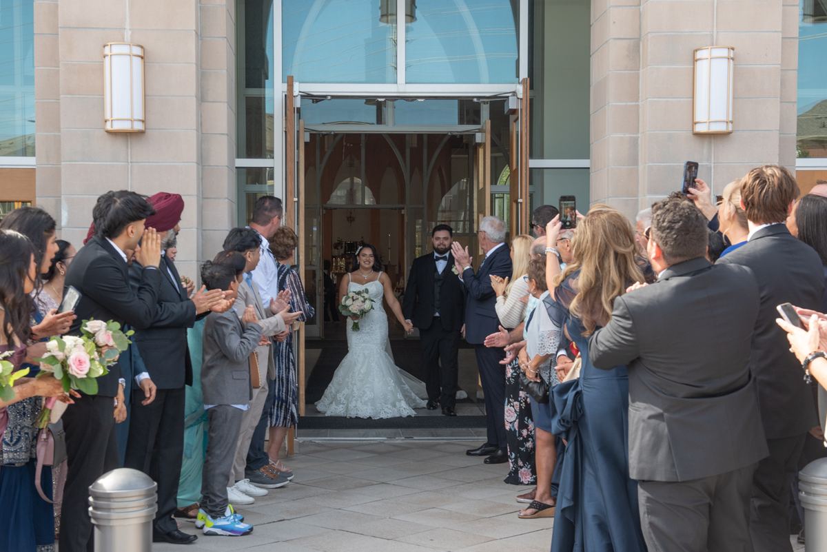 A bride and groom exit a modern church hand-in-hand, surrounded by applauding guests on either side.