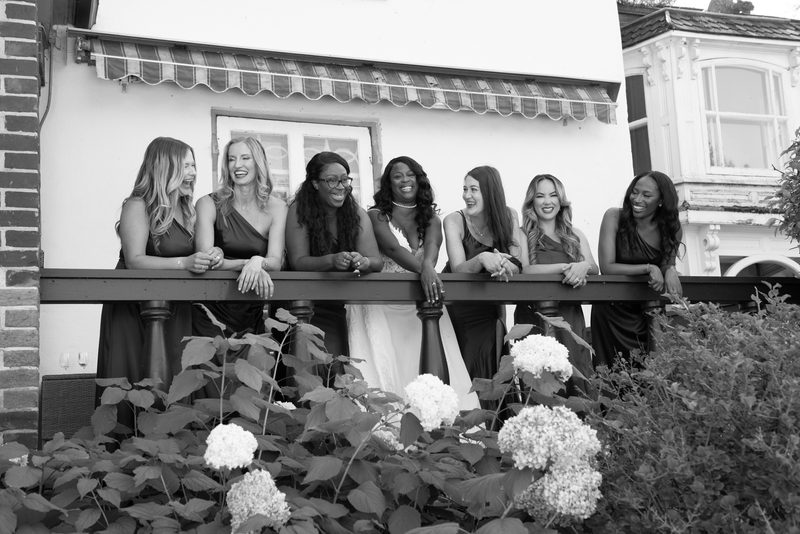 Bridesmaids in dark gowns laugh by a wooden railing, with a classic house and hydrangeas behind them.