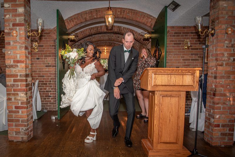 A smiling bride in a long white dress and her groom walk down a wooden aisle illuminated by hanging lanterns.
