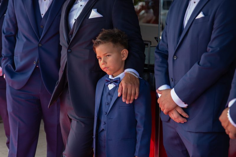 A young boy in a navy suit stands solemnly among adult men in matching attire at a wedding gathering.