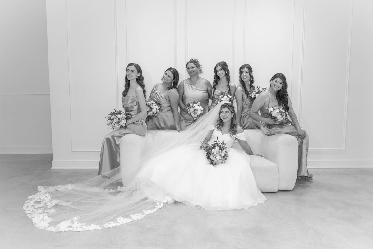 A bride and her bridesmaids pose joyfully in a minimalist studio with white walls, soft lighting, and floral bouquets.