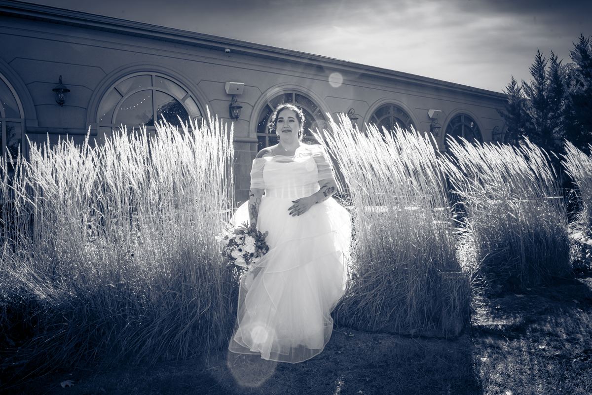 A bride in a white gown stands joyfully in a sunlit garden, framed by tall grasses and architecture.