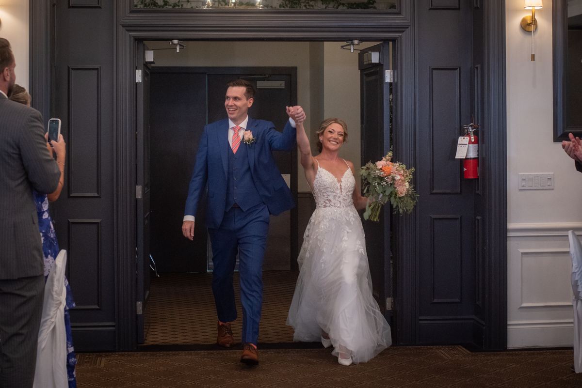 A smiling bride and groom hold hands as they exit through an ornate doorway, surrounded by guests celebrating their arrival.