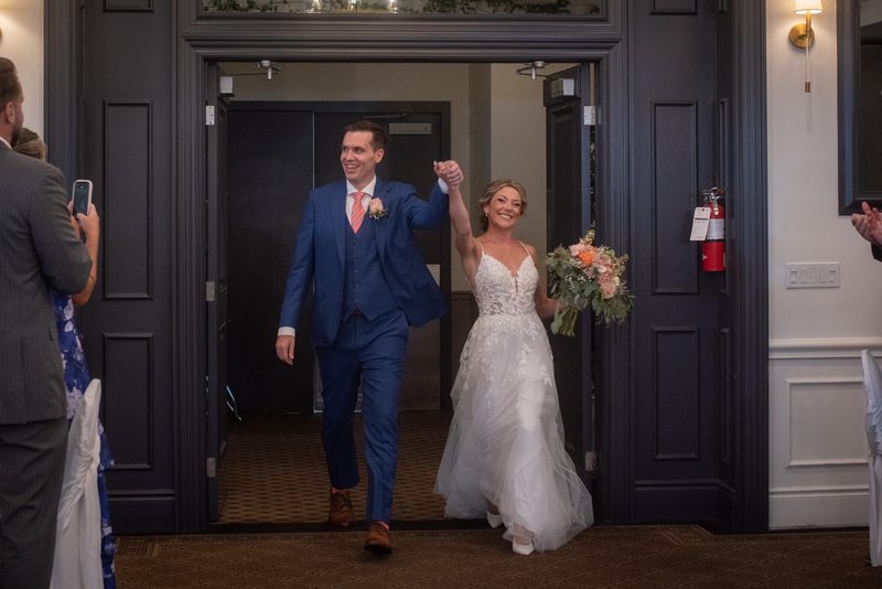 A smiling bride and groom hold hands as they exit through an ornate doorway, surrounded by guests celebrating their arrival.