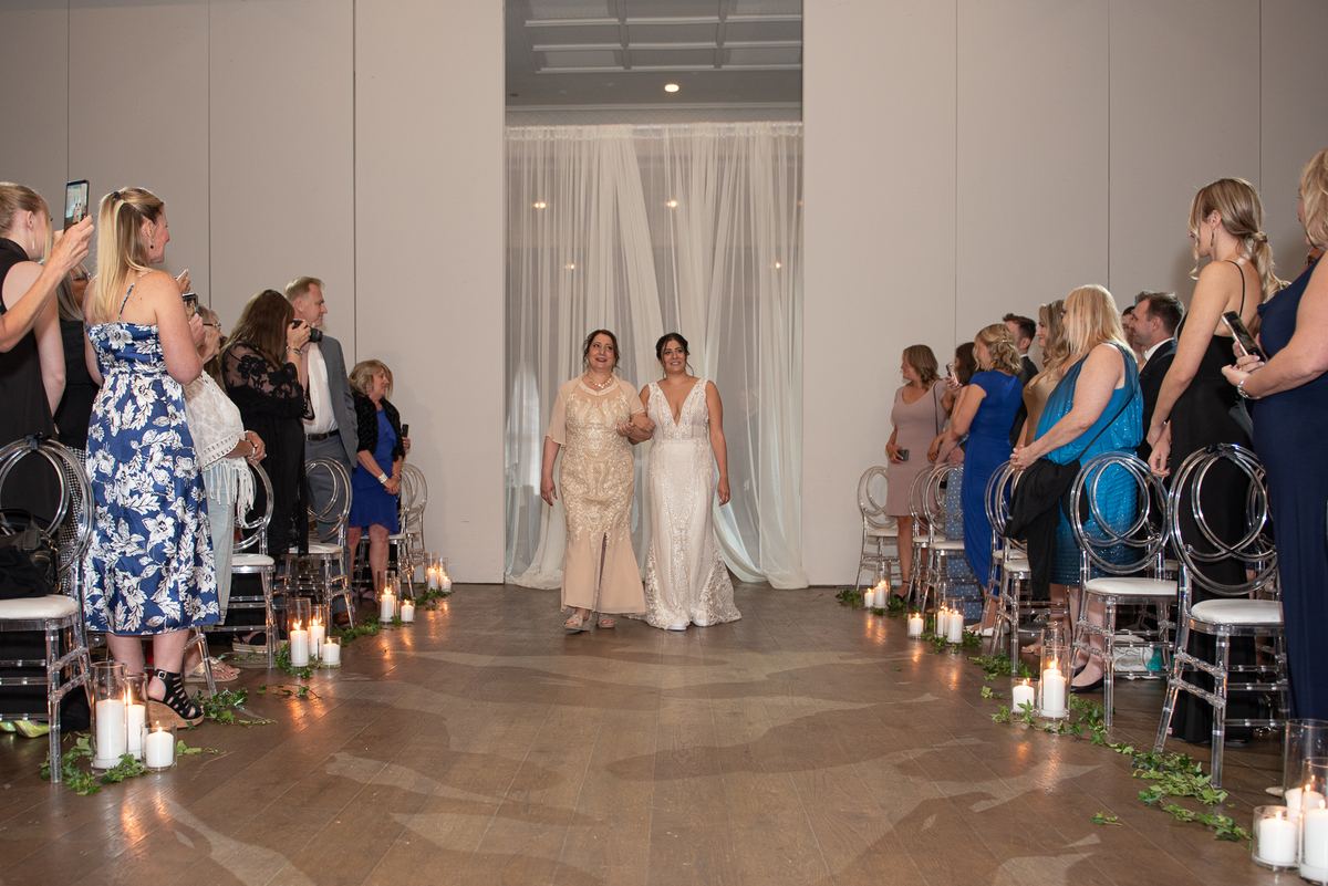 A bride in a white lace gown walks down a candlelit aisle toward a ceremony, framed by sheer white drapes.