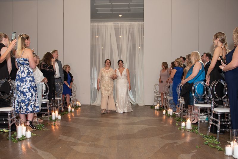 A bride in a white lace gown walks down a candlelit aisle toward a ceremony, framed by sheer white drapes.