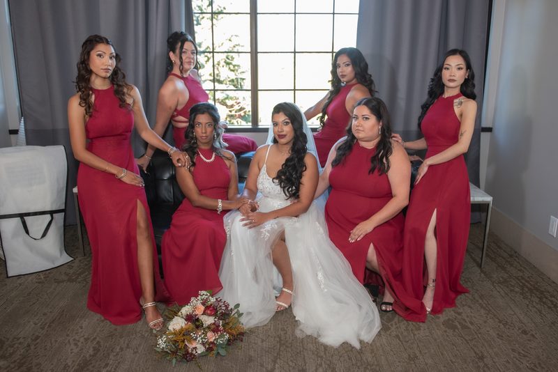 A bride and her bridesmaids pose in elegant crimson gowns, illuminated by natural light from large windows.