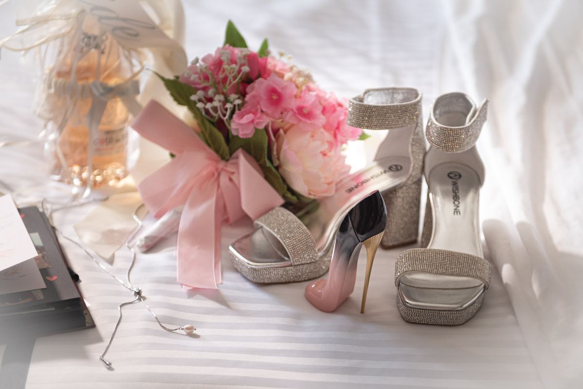 Sparkling high-heeled shoes, a delicate pink floral bouquet, and wedding accessories on a striped fabric in soft sunlight.