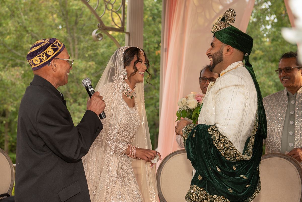 A bride in a white lehenga and veil laughs with the groom in an ornate sherwani as they exchange vows outdoors.