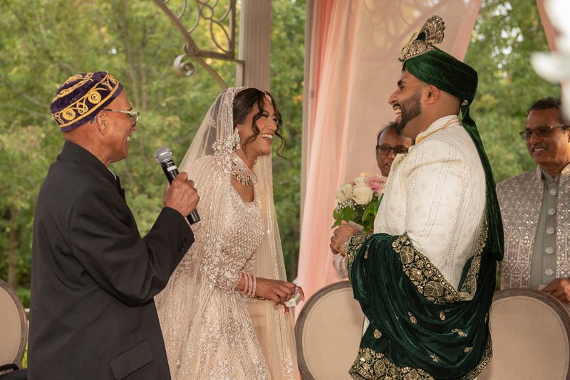 A bride in a white lehenga and veil laughs with the groom in an ornate sherwani as they exchange vows outdoors.