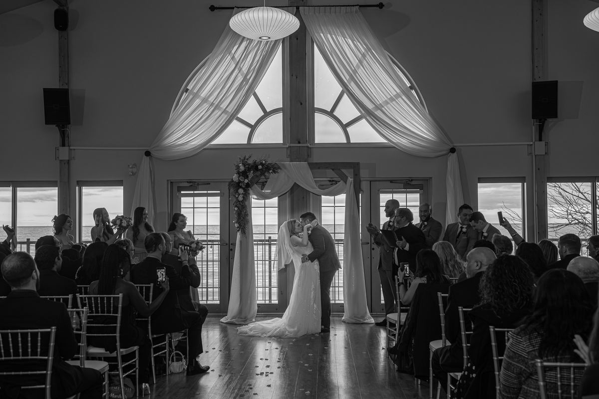 A bride and groom exchange a tender kiss at the altar, surrounded by draped fabric and an ocean view through a large window.