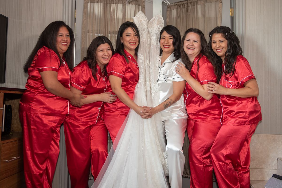 Bride in white satin robe and veil surrounded by smiling bridesmaids in red pajamas, holding hands in a circle.