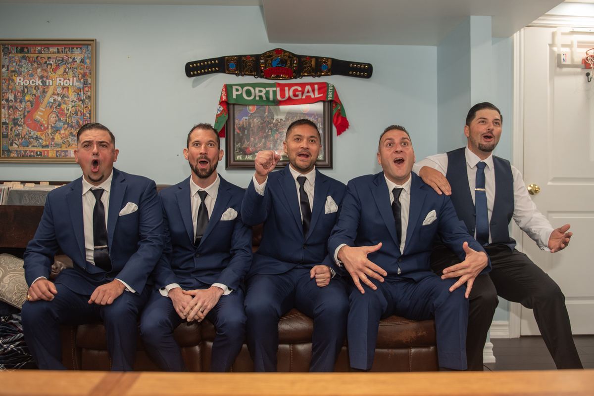 Five men in navy suits cheer exuberantly in a room decorated with Portugal memorabilia and a Rock n Roll poster.